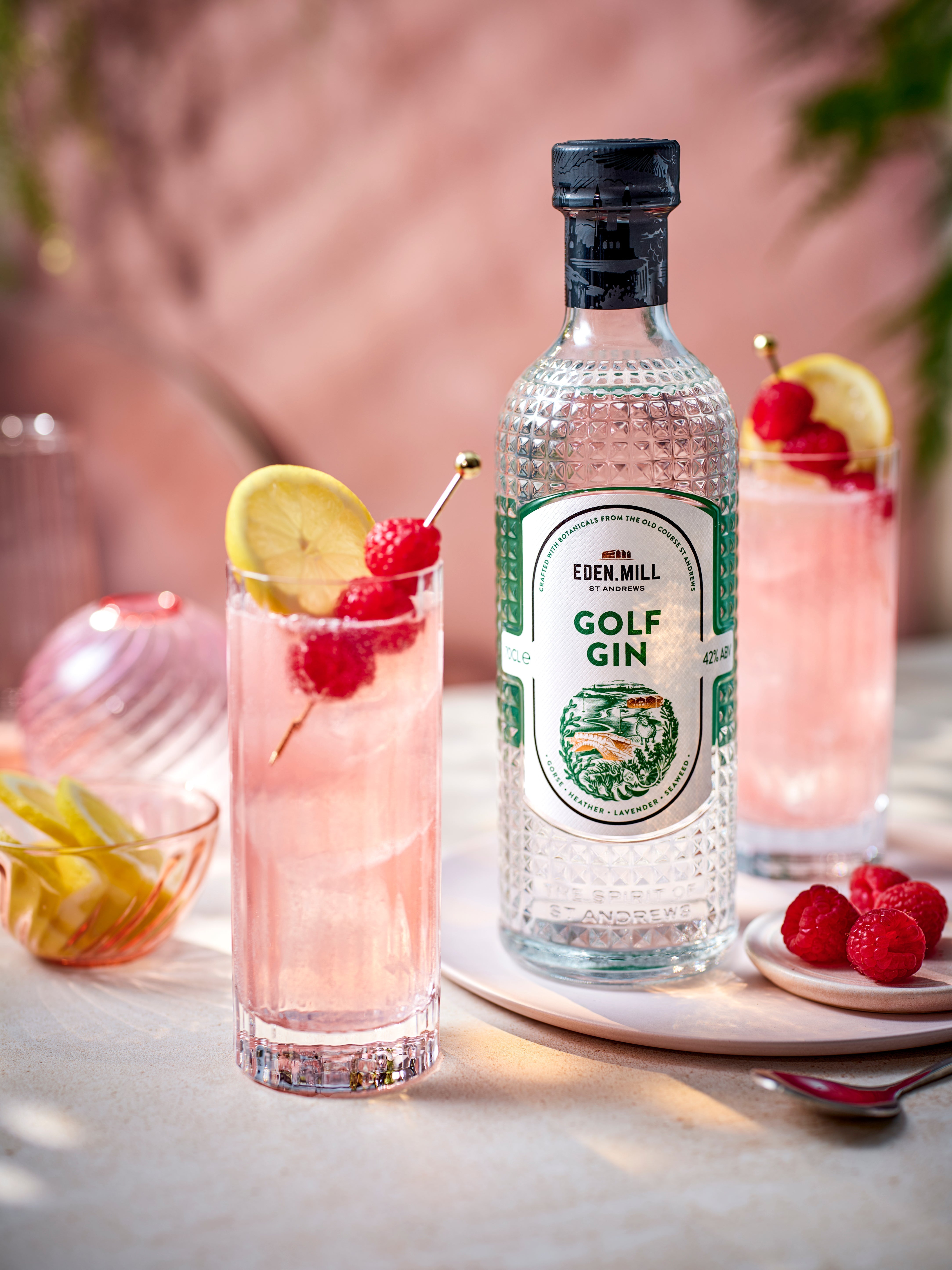 A picture of a bottle of Golf Gin next to a cocktail made with raspberry syrup and lemonade.
